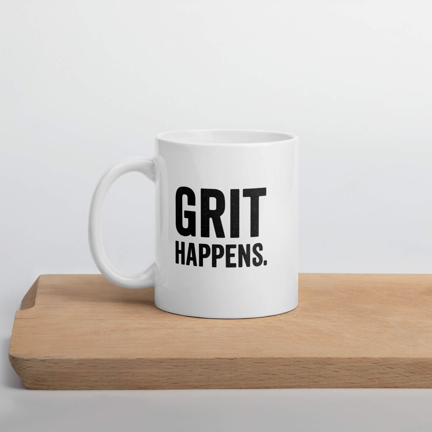 Front view of the Grit Collective "Grit Happens" 11oz white glossy ceramic mug on a wooden cutting board.