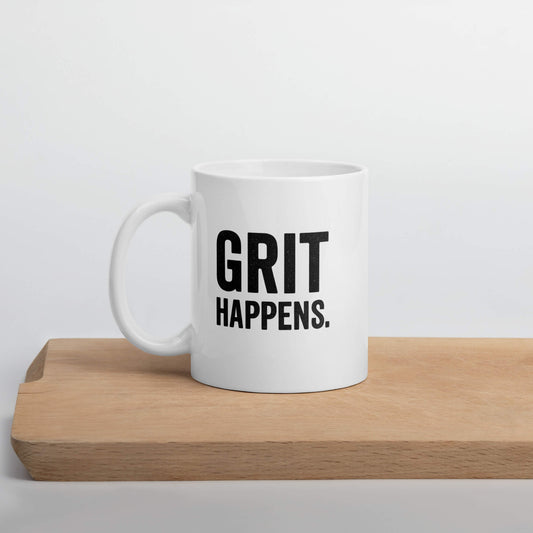Front view of the Grit Collective "Grit Happens" 11oz white glossy ceramic mug on a wooden cutting board.