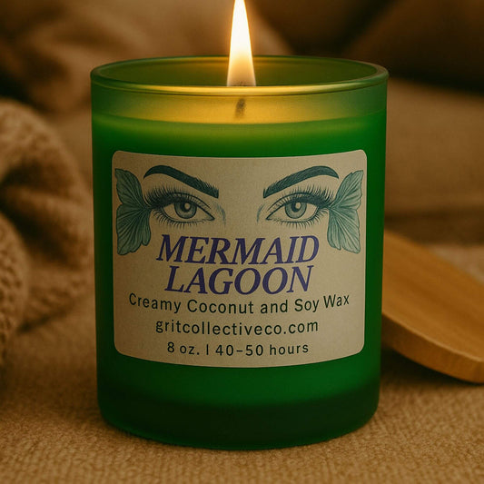 A lit "Mermaid Lagoon" creamy coconut and soy wax candle by Grit Collective Co. in a green glass jar with a bamboo lid, in a softly lit, cozy environment.