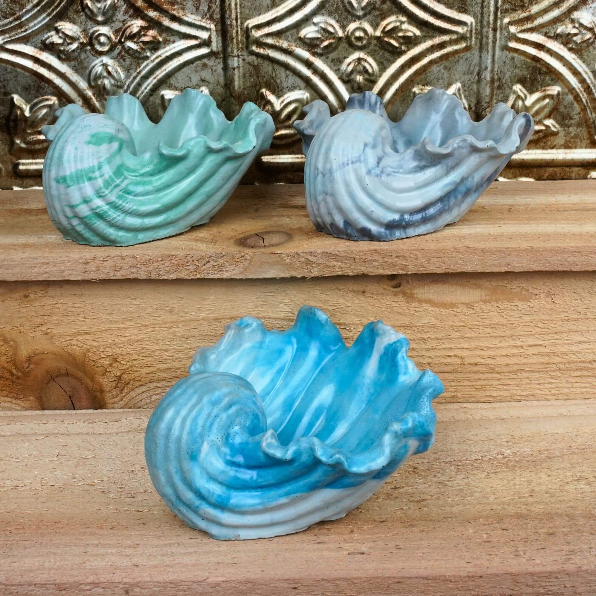 Set of three handcrafted concrete Ocean Bowls in blue, gray/lavender, and green colors, displayed on wooden shelves.
