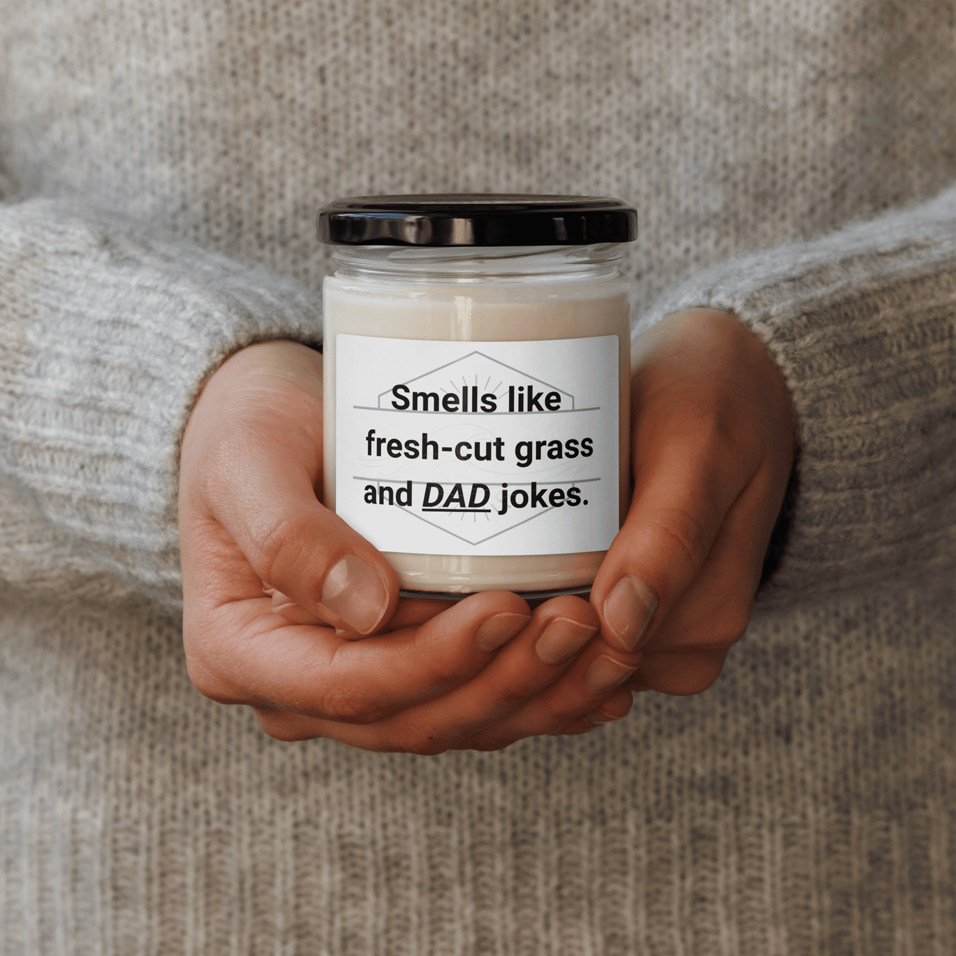 Hands holding a "Smells like fresh-cut grass and DAD jokes" 9oz soy wax candle.
