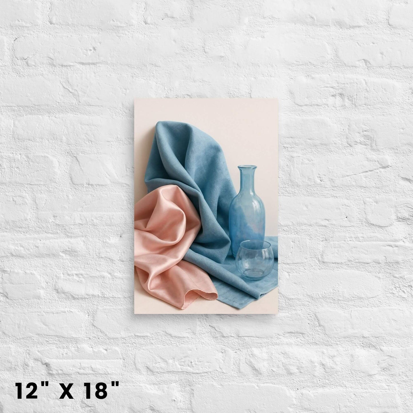 12"x18" canvas print of a still life with blue and pink fabric and glass items, mounted on a white brick wall.