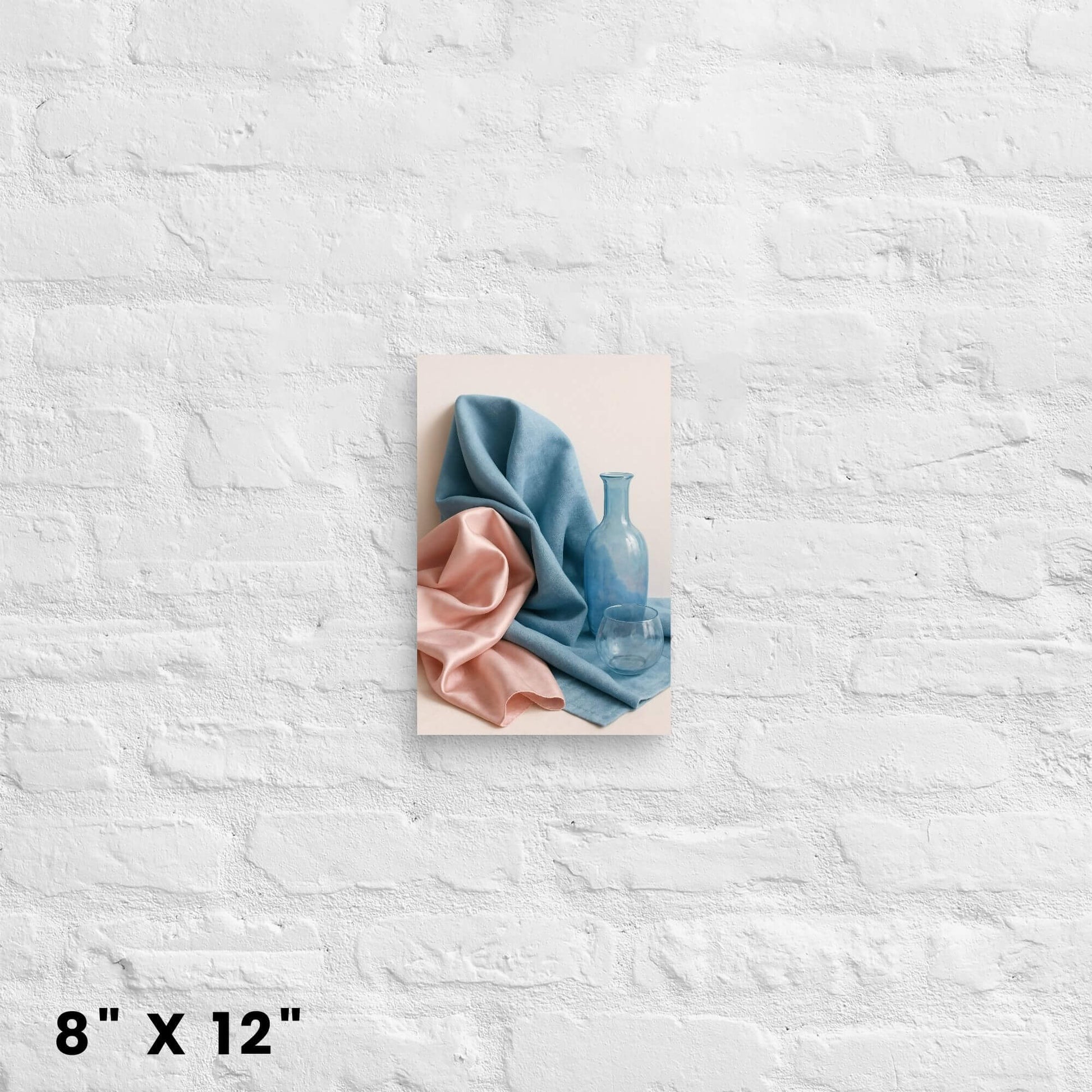 8"x12" canvas print showcasing a still life with draped blue and pink fabric, a blue bottle, and a glass, hanging on a white brick wall.