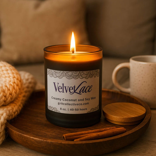 A lit Velvet Lace creamy coconut and soy wax candle by Grit Collective Co. in a black glass jar with a bamboo lid, resting on a wooden saucer with cinnamon sticks, a mug, and a cozy knit blanket in the background.