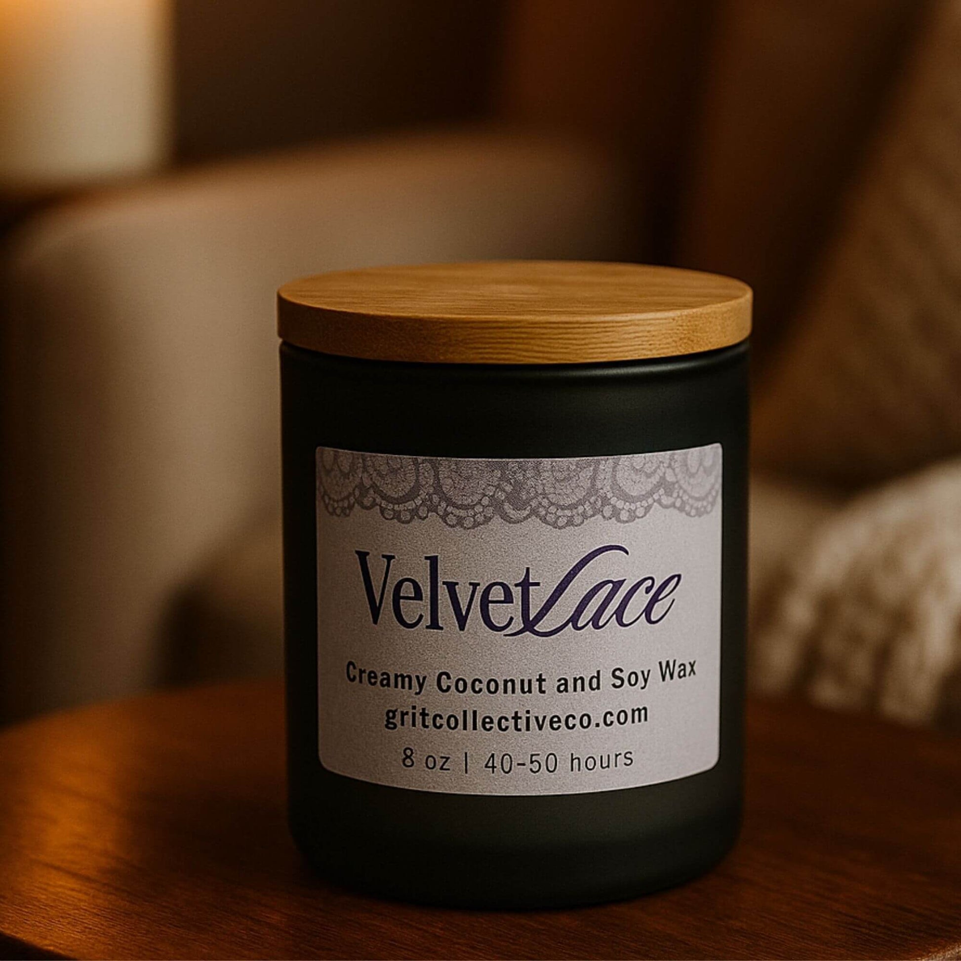 A Velvet Lace creamy coconut and soy wax candle by Grit Collective Co. in a black glass jar with a bamboo lid, sitting on a wooden table with a blurred, warm background.