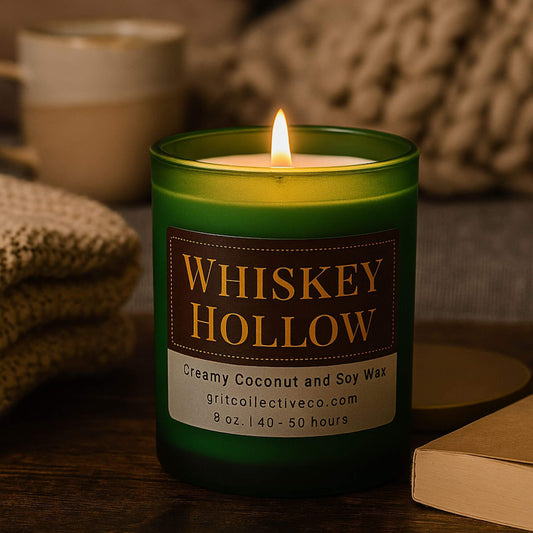 Whiskey Hollow" creamy coconut and soy wax candle by Grit Collective Co. in a green glass jar with a bamboo lid, placed on a wooden surface in a cozy setting.