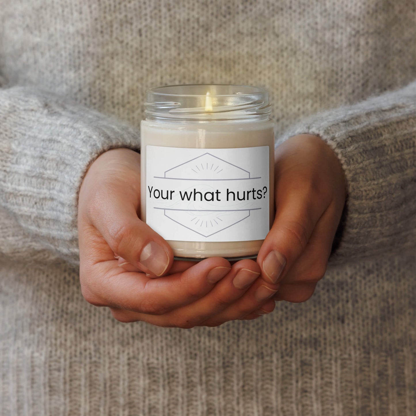 Hands holding a lit "Your what hurts?" funny scented soy candle, cozy lifestyle image.