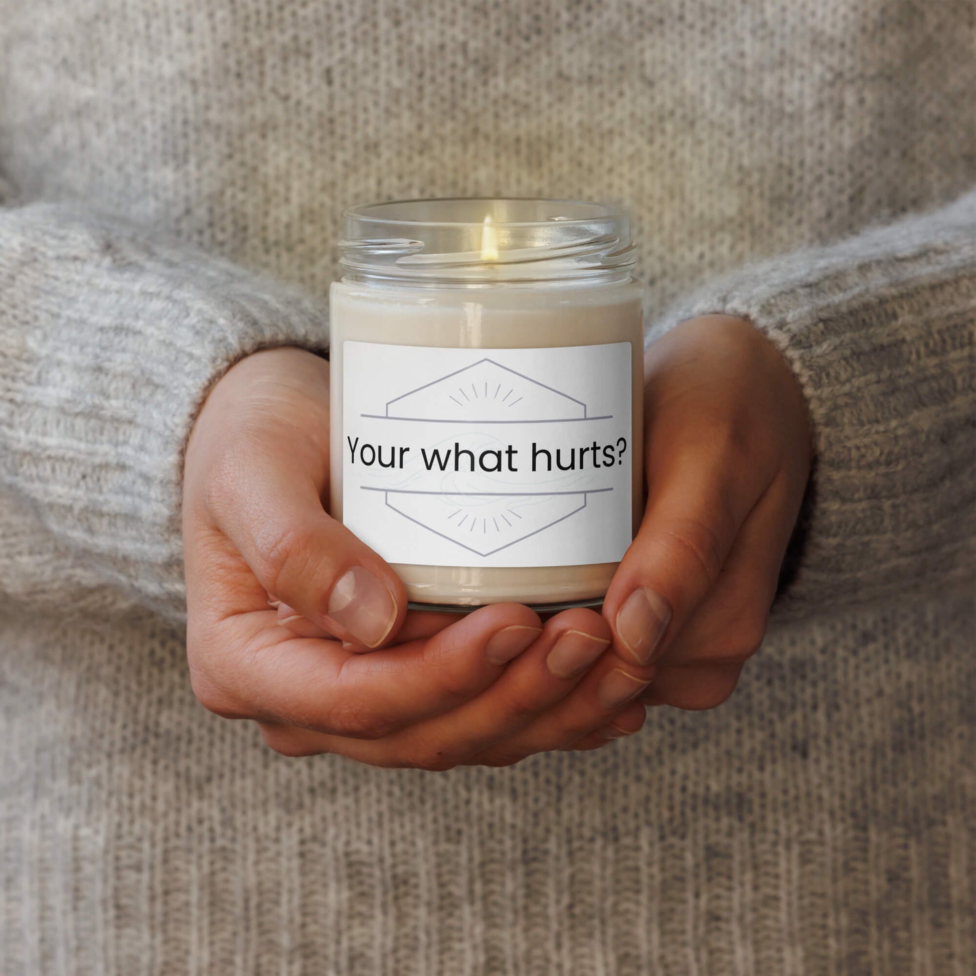 Hands holding a lit "Your what hurts?" funny scented soy candle, cozy lifestyle image.